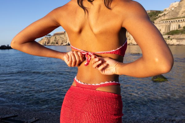 Dettaglio del copricostume rosso a crochet con decorazioni floreali rosa della linea Etna by Elisabeth Fashion, indossato in riva al mare.