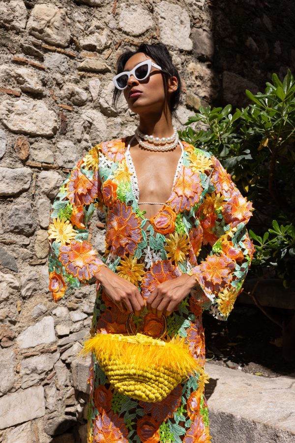 Dettaglio di una borsa gialla in corda con piume e manico dorato, accostata a un vestito floreale ricamato multicolore.