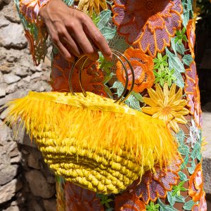 borsa gialla in corda con piume e manico dorato, accostata a un vestito floreale ricamato multicolore.