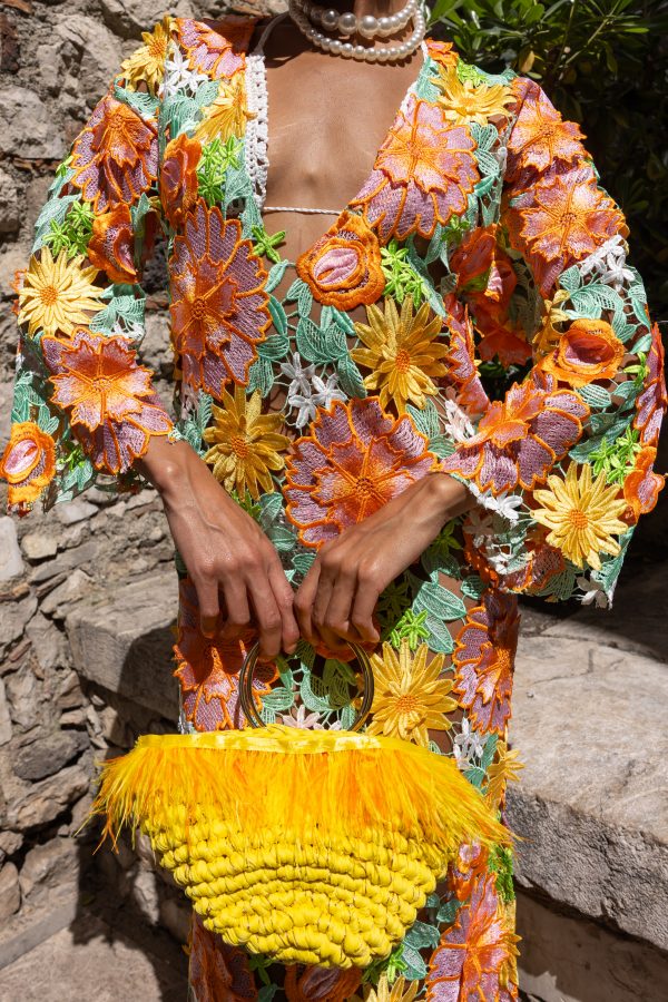 Dettaglio di una borsa gialla in corda con piume e manico dorato, accostata a un vestito floreale ricamato multicolore.