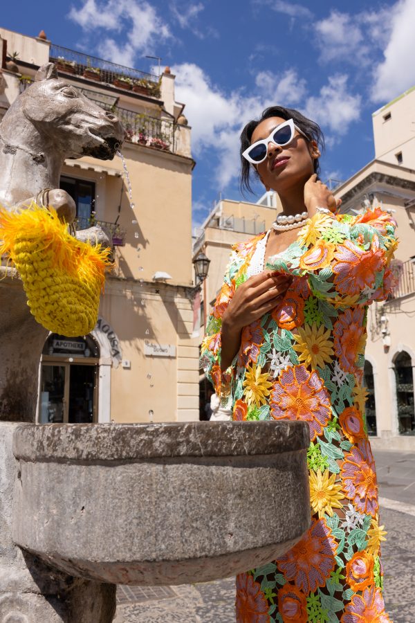 Dettaglio di una borsa gialla in corda con piume e manico dorato, accostata a un vestito floreale ricamato multicolore.