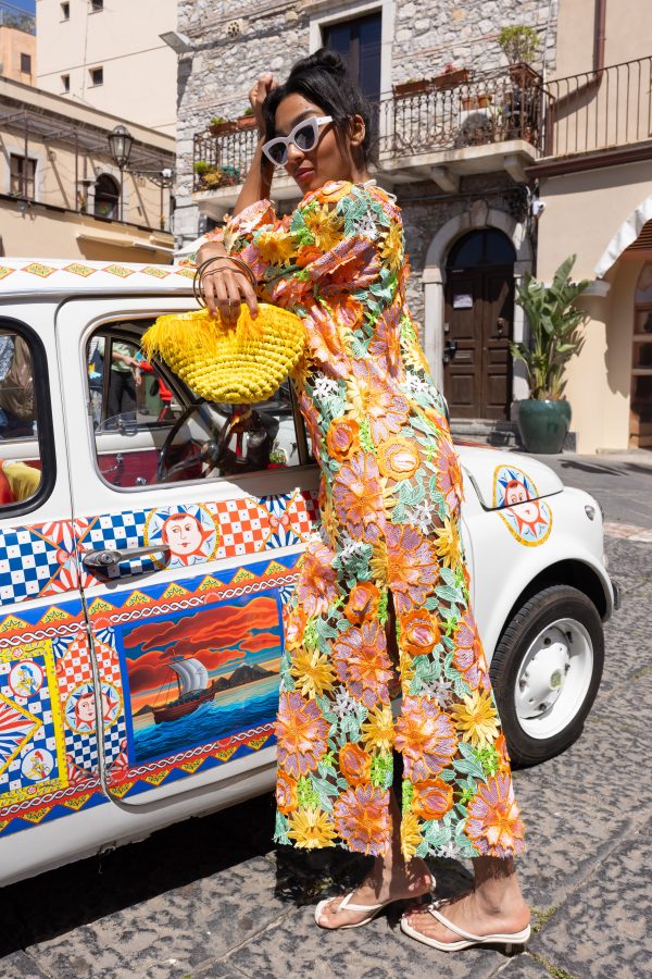 Dettaglio di una borsa gialla in corda con piume e manico dorato, accostata a un vestito floreale ricamato multicolore.