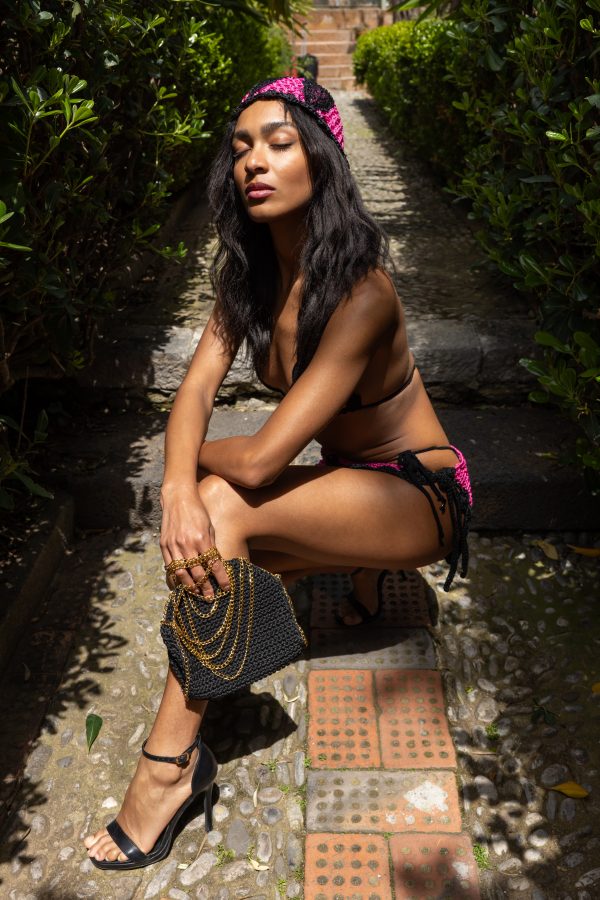 Borsa Vulcano Elisabeth Fashion, intrecciata a mano in fettuccia nera con catena dorata multi-strato, tenuta in mano da modella con outfit estivo elegante.